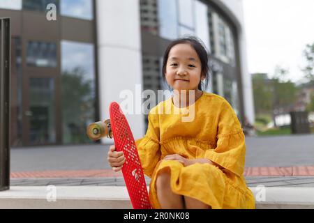 Glückliches asiatisches Mädchen mit Skateboard, das draußen in einer Stadt sitzt. Stockfoto