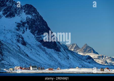 Schneebedeckte Berge erheben sich über dem Dorf Flakstad und dem Meer, Vareid, Lofoten, Nordland, Norwegen Stockfoto