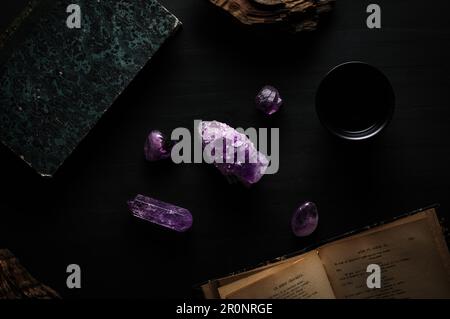 Amethyst-Edelstein-Detail auf schwarzem Tisch mit französischem Buch, Holzstück. Dunkle gotische Hexe, Hintergrund von oben Stockfoto