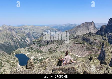 Eine Touristin sitzt auf einem Felsen hoch in den Bergen. Panoramablick auf eine große Bergkette und den See. Ergaki Naturpark, Krasnojarsk Region, Sibirien Stockfoto