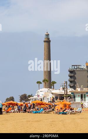Touristen am Strand unter orangefarbenen Sonnenschirmen mit dem Leuchtturm von Maspalomas an der Küste von Punta de Maspalomas. El Faro de Maspalomas. Las Palmas, Gr Stockfoto