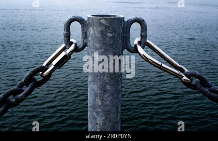 Metallpfosten auf einem Dock, mit rostigen Ketten und zwei silbernen oder verchromten Clips. Leicht gewelltes Flusswasser, Wasserrand. Dunkel, fast monochrom. Stockfoto