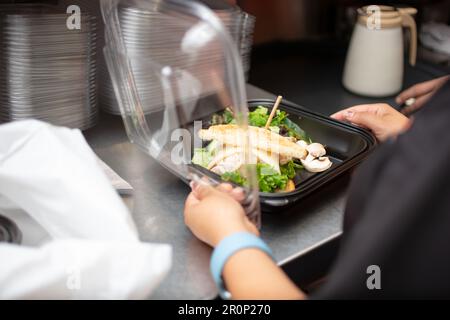 Ein Blick auf einen Mitarbeiter, der sich darauf vorbereitet, einen Lebensmittelbehälter für unterwegs in einer Restaurantküche zu verpacken. Stockfoto