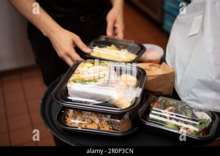 Ein Blick auf einen Mitarbeiter, der sich darauf vorbereitet, mehrere Lebensmittelbehälter für unterwegs in einer Restaurantküche zu verpacken. Stockfoto