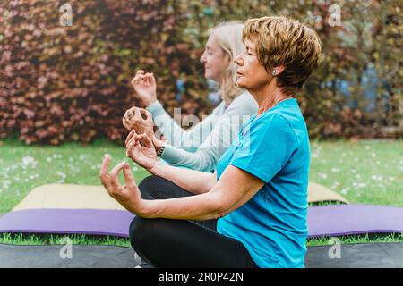 Eine Gruppe von Seniorinnen wird im Profil gesehen, sitzt in einem Garten auf Yogamatten und übt Meditation in der Lotusposition. Sie genießen alle einen Frieden Stockfoto