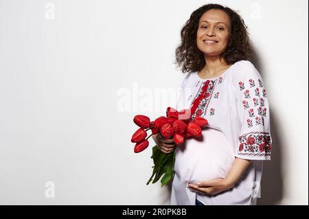 Entzückende Erwachsene schwangere Frau in einem bestickten ukrainischen Hemd, lächelnd, streichelnd, mit roten Tulpen posieren Stockfoto