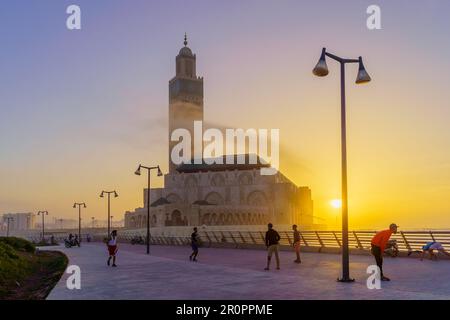 Casablanca, Marokko - 29. März 2023: Sonnenuntergangsszene der Hassan II Moschee mit Einheimischen, die Fußball spielen, Promenade und El Hank Leuchtturm in ca. Stockfoto