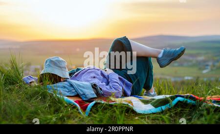 Eine Frau, die sich auf einem Hügel entspannt, mit einem Eimer, der das Gesicht bei Sonnenuntergang bedeckt Stockfoto