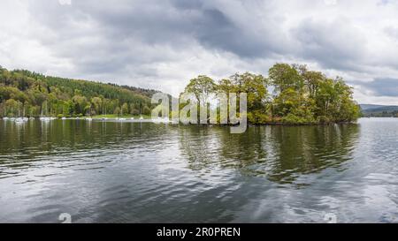 Eine kleine Insel, die nur von Bäumen bewohnt ist, die in einem mehrfarbigen Panorama vor der Mitchell Wyke Ferry Bay in Lake Windermere zu sehen sind. Stockfoto