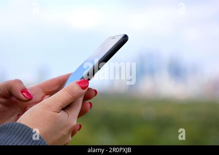 Weibliche Hände mit Smartphone Nahaufnahme auf unscharfen Wolkenkratzern. Eine Frau mit roter Maniküre, die im Park ein Handy benutzt Stockfoto