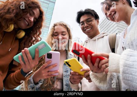Porträt von vier lächelnden Freunden, die von unten auf ihr Handy schauen. Junge Leute gehen wieder zur Schule. Stockfoto