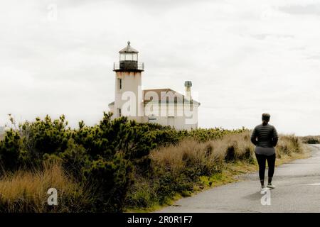 Der Leuchtturm des Coquille River in Bandon, Oregon, USA, im Bullards Beach State Park an der Küste von Oregon, PNW, mit einer Frau, die auf ein Gebäude mit hellen Bewölkungen zugeht Stockfoto