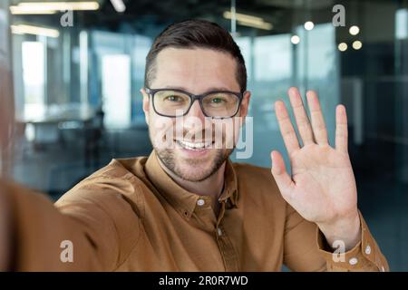 Erfolgreicher junger Geschäftsmann, der mit Freunden telefoniert, lächelt und winkt der Smartphone-Kamera, Arbeiter in der Pause. Stockfoto