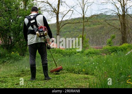 Ein Mann schneidet grünes Gras mit einem handgeführten Trimmer in seinem ländlichen Garten an einem Frühlingstag. Der Mann trägt Stiefel und konzentriert sich auf seine Arbeit Stockfoto