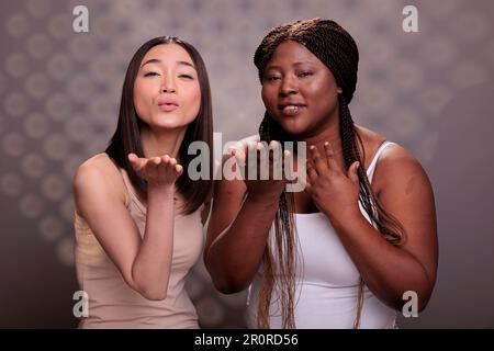 afroamerikanische und asiatische Frauen mit unterschiedlichen Körpertypen und ethnischer Zugehörigkeit, die einen Luftkuss schicken. Verschiedene Schönheitsmodelle, die Liebe und Zuneigung zeigen und im Studio posieren Stockfoto