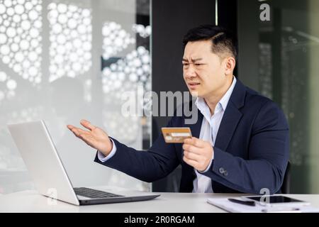 Frustrierter asiatischer Mann, der versuchte, Banktransaktionen und Einkäufe im Online-Shop zu tätigen, ein Geschäftsmann betrog und traurig, als er im Büro mit einem Laptop und einer Bankkreditkarte arbeitete. Stockfoto