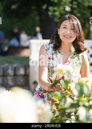 Eine weiße Frau bewundert blühende Rosen im öffentlichen Park. Sommerliche Stimmung. Tropische Pflanzen und Blumen blühen im Garten. Stockfoto