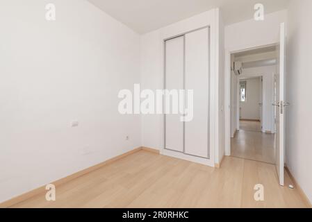 Ein leeres Zimmer in einem Apartment mit eingebautem Kleiderschrank mit weißen Schiebetüren, hellem Holzfußboden und weißen Zugangstüren Stockfoto