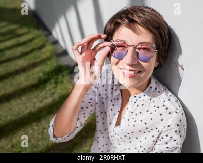 Lächelnde Frau in farbenfroher Sonnenbrille ruht sich auf dem Rasen im Stadtpark aus. Natur in der Stadt. Entspannen Sie sich nach der Arbeit im Freien. Sommerliche Stimmung. Stockfoto