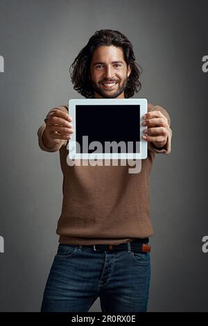 Du hast es hier zuerst gesehen. Studiofoto eines gutaussehenden jungen Mannes, der ein digitales Tablet mit einem leeren Bildschirm vor einem grauen Hintergrund hält. Stockfoto