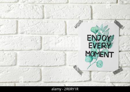 Karte mit Text Genießen Sie jeden Moment an der weißen Backsteinwand, Platz für Text Stockfoto
