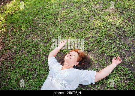 Reife hispanische Frau, die auf dem Gras liegt, mit ihren Armen in einer entspannten Position lächelt und einen Moment der Ruhe genießt. Stockfoto