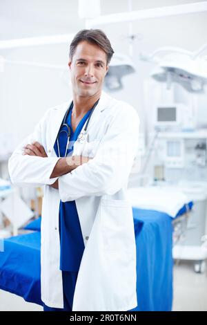 Der Arzt, auf den Sie hoffen. Ein glücklicher männlicher Arzt, der im Operationssaal mit gekreuzten Armen steht. Stockfoto