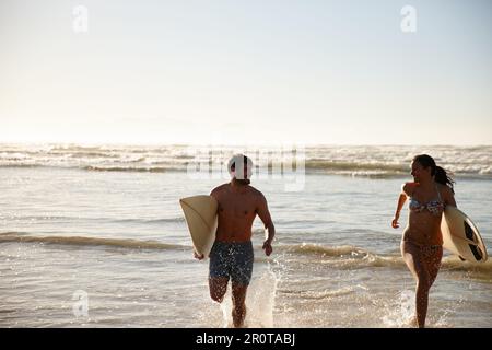 Das Beste aus dem Sommer machen. Ein junges Paar, das mit seinen Surfbrettern aus dem Meer rennt. Stockfoto