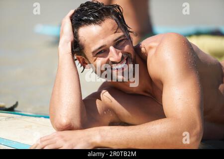 Das ist das Leben für mich. Ein junger Mann, der sich entspannt und lächelt, während er auf seinem Surfbrett im Sand liegt. Stockfoto