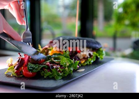 Gabel und Messer essen Salat in einem Restaurant köstliches leckeres Mittagessen Abendessen Mittagessen Entspannung herrliche Speisen Burger in schwarzem Tintenfisch Brötchen wundervoll servierend Stockfoto