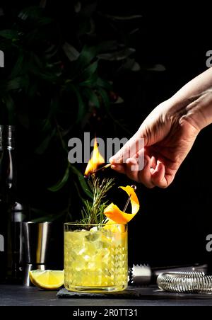 Penicillin-Cocktailgetränk mit Scotch, Whiskey, Sirup, Zitronensaft und Eis in Glas mit Orangenschale. Der Barkeeper zündet Rosmarin an. Schwarzer Hintergrund Stockfoto