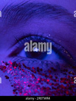Extreme Nahaufnahme des weiblichen Auges mit glänzendem Make-up mit Funkeln. Kinoähnliche Makroaufnahme eines weiblichen Augen beleuchteten rosa-blauen Neonlichts Stockfoto