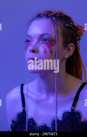 Nachtporträt mit Frauengesicht in Kinoqualität mit glänzendem Bühnenschmink und Pailletten, leuchtendem pinkblauem Neonlicht. Studiofoto eines jungen Weibchens Stockfoto