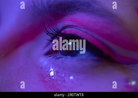 Extreme Nahaufnahme des weiblichen Auges mit glänzendem Make-up mit Funkeln und Pfeilen. Kinematische Makroaufnahme des weiblichen braunen Auges im Neonlicht Stockfoto