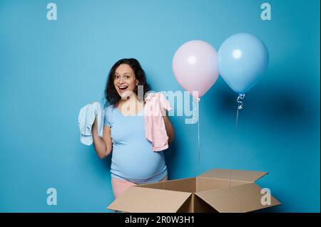 Glückliche schwangere Frau, die einen blau-pinkfarbenen Neugeborenen-Body zeigt, isoliert auf blauem Hintergrund. Das Partykonzept der Geschlechter Stockfoto
