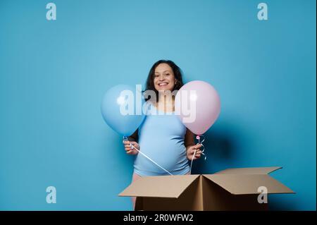 Wunderschöne schwangere Frau, die blaue und rosa Ballons hält, lächelt in die Kamera, isoliert auf blauem Hintergrund Stockfoto