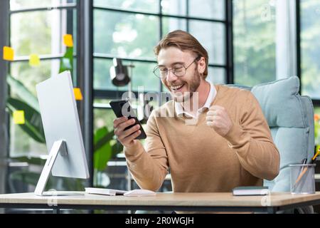 Ein junger Mann arbeitet im Büro an einem Tisch mit einem Laptop. Er hält das Telefon in den Händen, sieht glücklich und schockiert auf den Bildschirm, hob seine Brille auf sein Gesicht, um überrascht zu sein. Ich habe gute Neuigkeiten. Stockfoto
