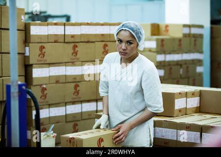 Kostanay, Kasachstan - 14. Mai 2012: Moderne Süßwarenfabrik Bayan-Sulu. Junge asiatische Arbeiterin im Lager. Pappkartons auf dem Hintergrund. Stockfoto