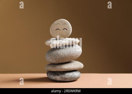 Steinstapel mit gezeichnetem fröhlichem Gesicht auf dem Tisch vor dunkelbeigem Hintergrund. Zen-Konzept Stockfoto