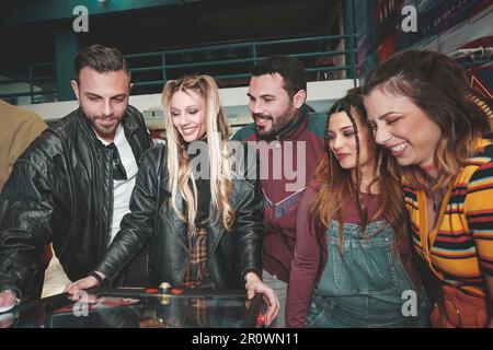 Eine junge blonde Weiße mit Sommersprossen im Gesicht spielt Flipper in einer Spielhalle, während ihre Freunde ihr zuschauen und sie anfeuern. Sie genießen einen Stockfoto