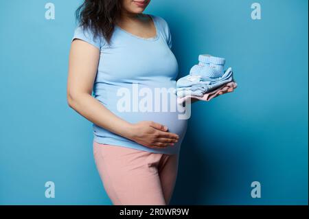 Nahaufnahme schwangere Frau, die Hand auf den Bauch legt, mit Neugeborenen Kleidung und Babystiefeln posiert, isolierter blauer Hintergrund Stockfoto