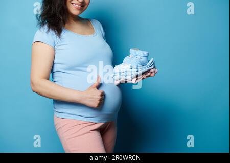 Nahaufnahme einer schwangeren Frau mit großem Bauch, zeigt Daumen nach oben, lächelt vor der Kamera, hält Neugeborenen-Kleidung in der Hand, isoliert auf blau Stockfoto