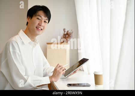 Ein kluger und attraktiver junger asiatischer Freiberufler oder Student im weißen Hemd sitzt mit seinem tragbaren Tablet in einem minimalistischen Café. Stockfoto