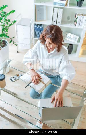 Frau im Büro am Computer. Sie trägt ein weißes Hemd und blaue Jeans, die an einem Tisch sitzen, auf dem ein Laptop und ein Notizblock stehen. Funktioniert in Stockfoto