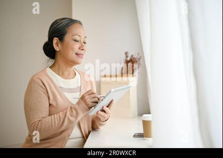 Charmante und glückliche, reife 60s-asiatische Frau in legerer Kleidung, die ihr digitales Tablet in einem Café verwendet. Stockfoto