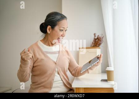 Aufgeregte und überraschte asiatische Seniorin sitzt am Fenster, schaut auf ihren Tablet-Bildschirm, schreit vor Freude, jubelt, feiert gute Nachrichten, gewinnt Stockfoto