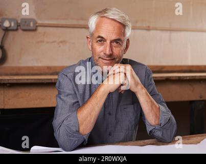 Stolz auf seine Werkstatt. Porträt eines Seniorenmannes, der an seinem Schreibtisch in seiner Werkstatt sitzt. Stockfoto