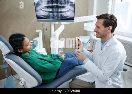 Lächelndes männliches Zahnarzthalter-Zahnmodell während der Beratung eines kindlichen Patienten in der Zahnklinik, Kopierraum Stockfoto