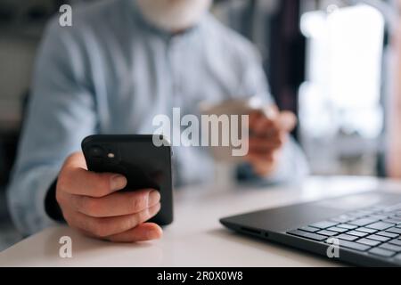 Nahaufnahme eines unbekannten, bärtigen Geschäftsmanns, der an einem Laptop arbeitet, ein Smartphone benutzt, eine Tasse Kaffee hält, sitzt Stockfoto