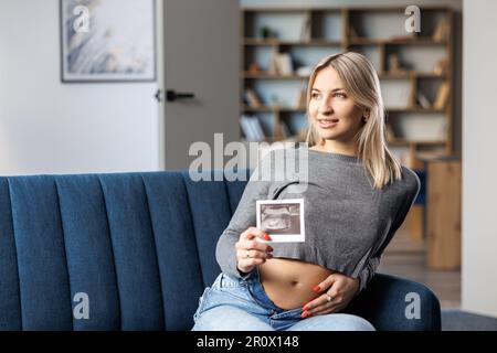 Foto einer schwangeren Frau zu Hause, die auf dem Sofa neben einem wunderschönen Blumenstrauß sitzt und in der Hand den Ultraschall-Scan-Druck des Babys hält Stockfoto